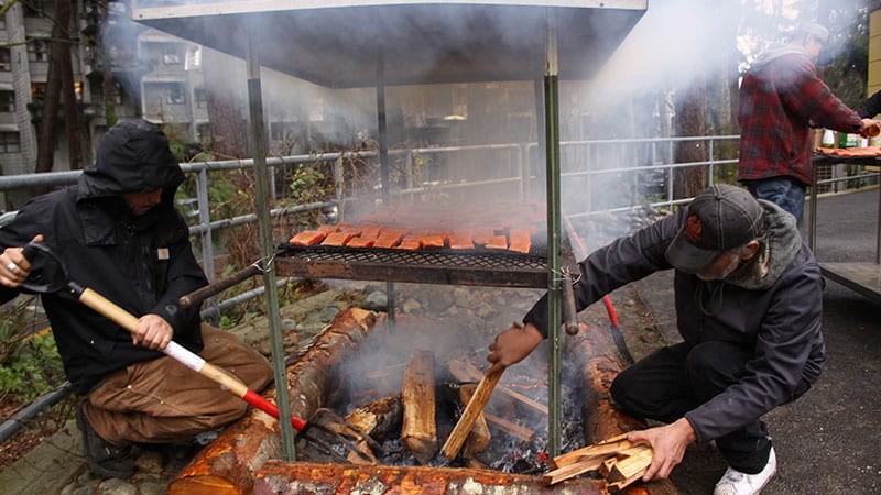 Members of the Muckleshoot Tribe cook wild salmon in a traditional preparation over a fire pit at a 2017 community gathering to learn about the effects of GE salmon. Photo credit: Johanna Lundahl photography.